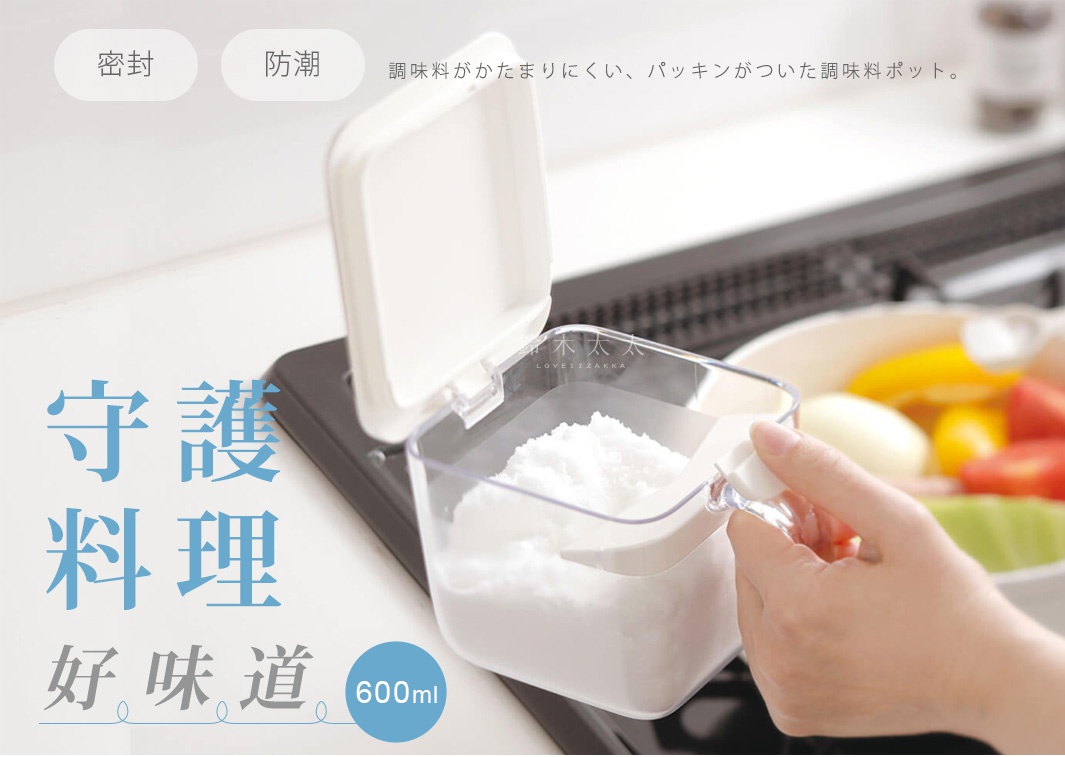這張圖片展示了一款容量為600毫升的透明方形調味料盒，盒蓋採用白色設計並配有密封膠條以達到防潮效果。畫面中一隻手正拿著調味料盒內的白色塑膠量匙，盒身內部裝有白色調味料。背景為廚房爐台，整體呈現出簡潔實用的家居生活氛圍。圖片中標示了密封、防潮及調味料盒說明文字，強調能防止調味料結塊。