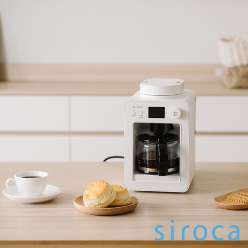 白色 SC-A3520 自動研磨咖啡機，配有玻璃咖啡壺，上方有黑色操作面板和旋鈕。桌面上擺放著一杯咖啡、兩個司康餅和幾塊餅乾。