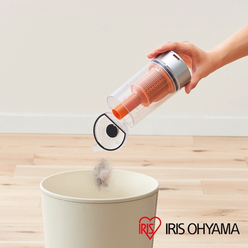 手持吸塵器集塵筒傾斜，將灰塵倒入米白色垃圾桶，集塵筒為透明塑膠材質，內有橘色濾網與吸管。