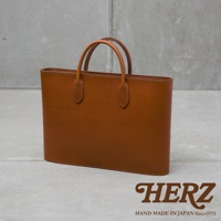 職人手作皮革Organ薄型公事包，採用光滑棕色皮革製成，配有圓潤的提把和精緻的縫線細節，整體設計簡潔大方。