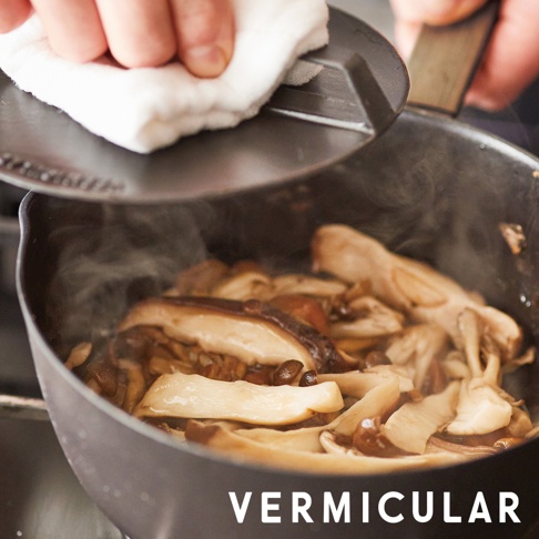 一雙手正隔著白色抹布，將一個黑色琺瑯鑄鐵落蓋蓋在正在烹煮菇類食材的 VERMICULAR 鑄鐵雪平鍋上，鍋內冒出陣陣熱氣。