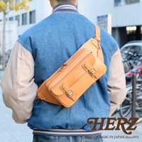 棕色皮革製作的斜背包，斜挎於身穿牛仔外套的男士背部，包身有兩個翻蓋口袋，均以雙釦設計固定，展現職人手作質感。