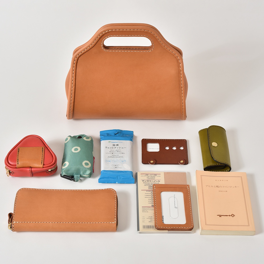 淺棕色皮革拱型手袋，搭配多種配件，包括紅色三角形零錢包、綠色圓點環保袋、濕紙巾、棕色卡夾、綠色鑰匙包、長款拉鍊錢包、卡片夾及日文書籍。