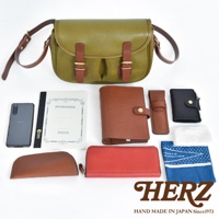 橄欖綠皮革傳統釣魚包，搭配棕色皮革翻蓋與束帶，附有金屬扣。周圍擺放著棕色皮夾、紅色長夾、黑色小錢包、筆記本、手機、手機套及印有藍白圖案的布巾。