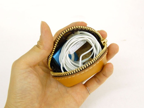 手持一個棕色皮革製成的貝殼造型零錢包，拉鍊開口露出內部收納的白色耳機和藍色小物品。