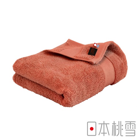 一條珊瑚色今治匹馬棉浴巾，質地柔軟，邊緣處有黑色品牌標籤和紅藍條紋裝飾。