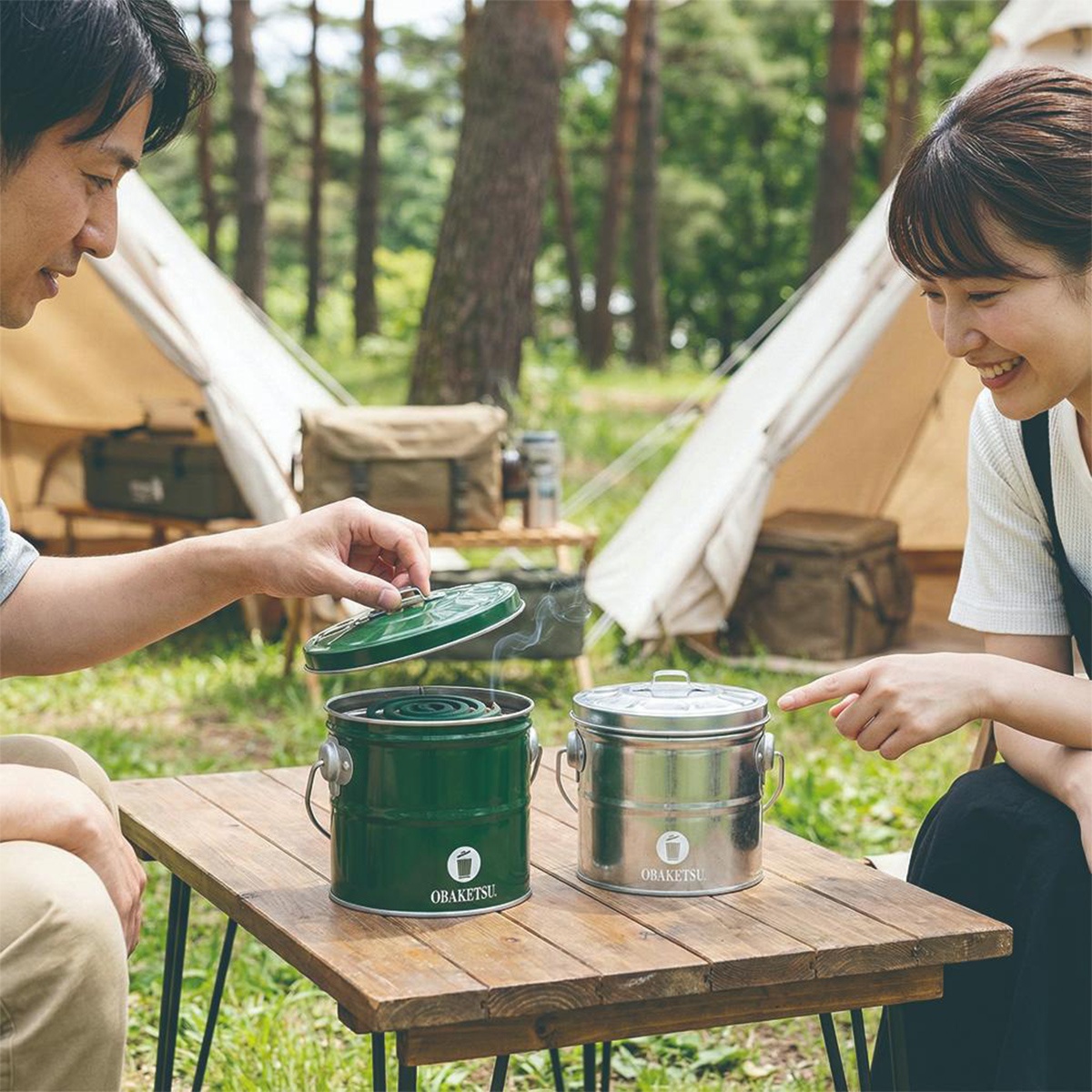 一對男女在戶外野營，男子正打開綠色OBAKETSU蚊香桶，旁邊擺放著銀色同款蚊香桶。