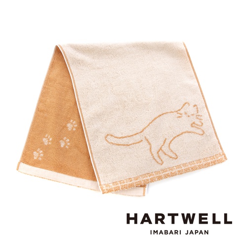 這是一條Hartwell今治製的雙色毛巾，設計呈現柔和的米色與棕色調，米色面上勾勒著一隻慵懶貓咪的簡約線條，而棕色面上則點綴著可愛的貓掌印記，毛巾下方並印有HARTWELL IMABARI JAPAN品牌字樣。