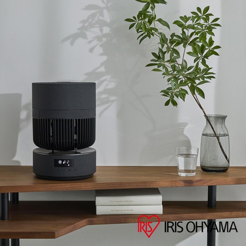 一台深灰色的IRIS OHYAMA 360度美型空氣循環扇放置在木質層架上，造型簡約優雅，下方展示螢幕顯示運作數值，旁側配有一杯水及插在玻璃花瓶中的綠色枝葉，層架下方擺放著兩本白底書籍。