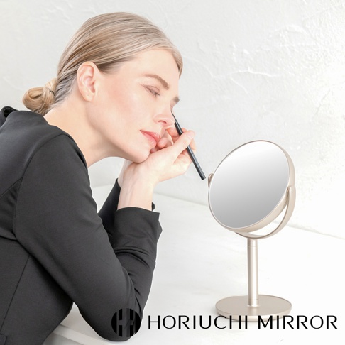 香檳金色的桌上型化妝圓鏡，鏡面向上傾斜，附有圓形底座和支架。一位身穿黑色上衣的金髮女子正側身使用細長筆狀物描畫眉毛。