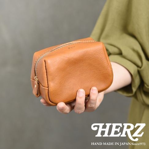 一位身穿綠色上衣的人手持一個淺棕色皮革拉鍊收納包，該包款外觀簡約並印有Organ字樣，照片右下角帶有HERZ品牌標誌及HAND MADE IN JAPAN Since1973字樣，展現日式職人手作皮革製品的質感。