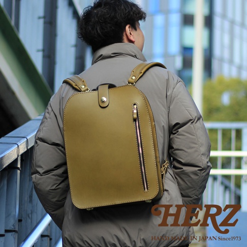 橄欖綠皮革製成，方形簡約設計的兩用後背包，搭配銀色拉鍊和金屬扣環，展示於穿著灰色外套的男性背後。