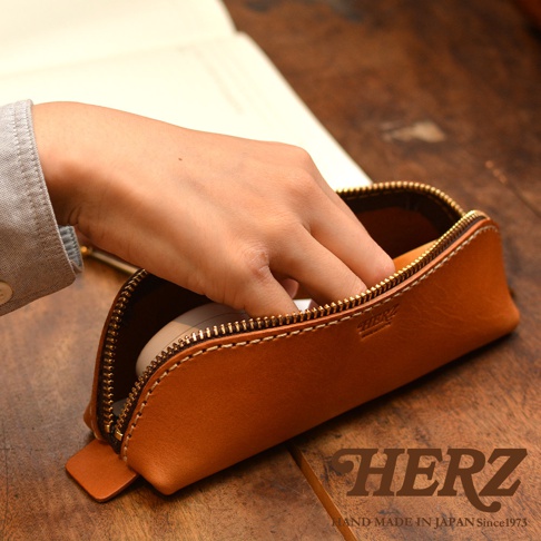 棕褐色皮革製成的三角形收納筆袋，搭配金色拉鍊，手工縫線，置於木質桌面。