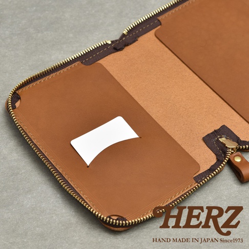 HERZ 品牌標誌，下方為 HAND MADE IN JAPAN Since1973 字樣，整體為棕色皮革收納包，內有卡槽，拉鍊為金色。