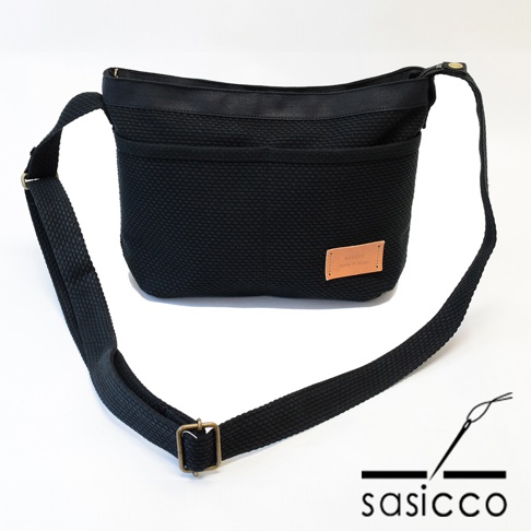 黑色三河木綿刺子織斜背包，附有可調節織帶和皮革品牌標籤，設計簡約實用。