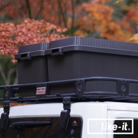 兩個黑色可疊式波浪紋收納箱，其中一個箱子上面印有「SMITTYBILT DEFENDER ROCK」標誌，擺放在汽車頂部的黑色置物架上，背景為模糊的秋季樹葉。