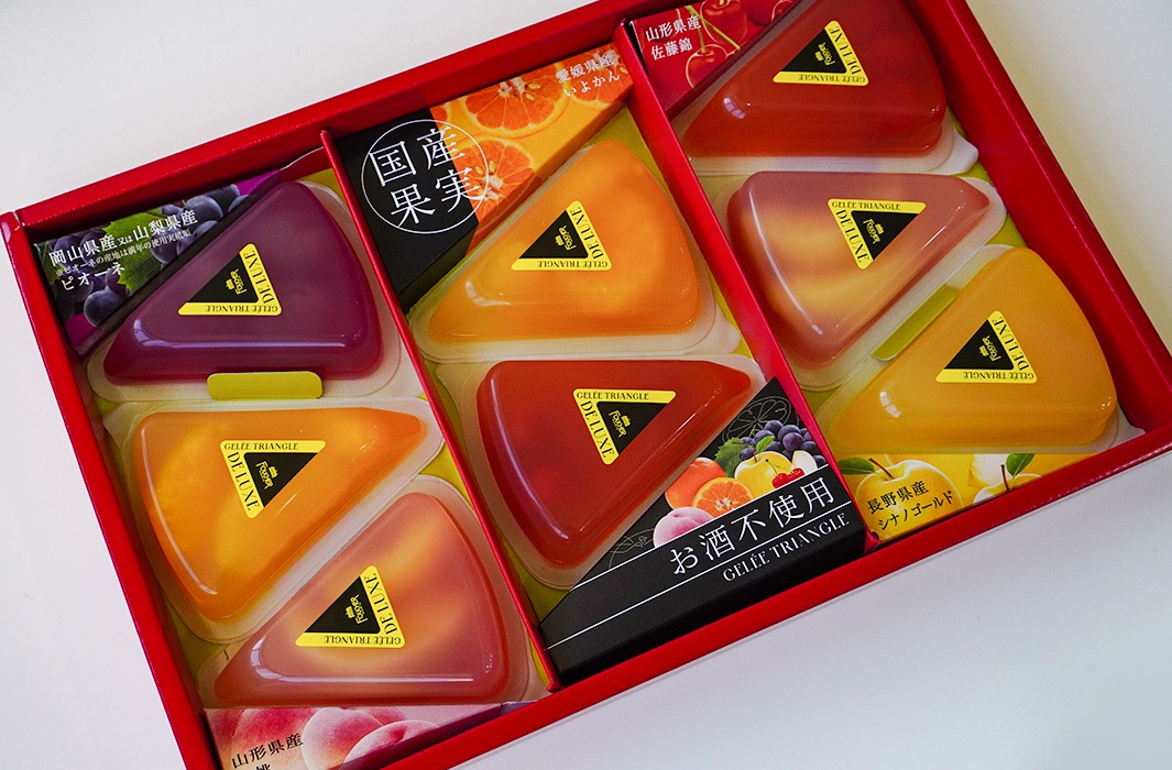 紅色禮盒內盛裝著八個三角形果凍，顏色包含紫色、橘色、紅色和黃色。果凍表面貼有黃色三角形標籤，標示著「GELEE TRIANGLE DELUXE」。部分果凍旁有印製水果圖案的紙盒，如葡萄、橘子、櫻桃、桃子和蘋果。