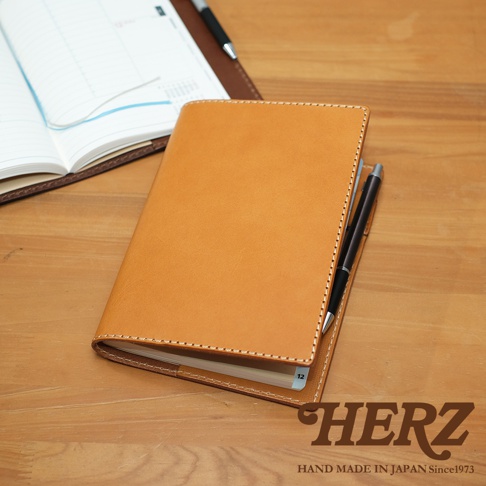 職人手作皮革B6記事本書套，展示卡其色皮革材質，車縫線精緻，內含一本記事本，並有一支黑色鋼筆夾在書套側邊。