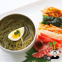 一碗裝有海苔蕎麥麵的銀色碗，麵條呈綠色，上面放有半顆水煮蛋和嫩綠的芽菜。旁邊的白色盤子上擺放著多種配料，包括韓式泡菜、深綠色蔬菜、豆芽、蟹肉棒和章魚腳。