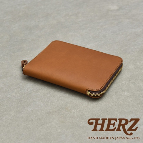 HERZ 品牌標誌，下方為 HAND MADE IN JAPAN Since1973 字樣，圖片主體為一個棕色皮革拉鍊式記事本收納包，置於灰色紋理背景上。