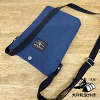 深藍色刺子織紋理平板收納側背包，配有黑色織帶和金屬釦件，背包上有黑色與紅色相間的品牌標籤，整體置於木紋背景上。