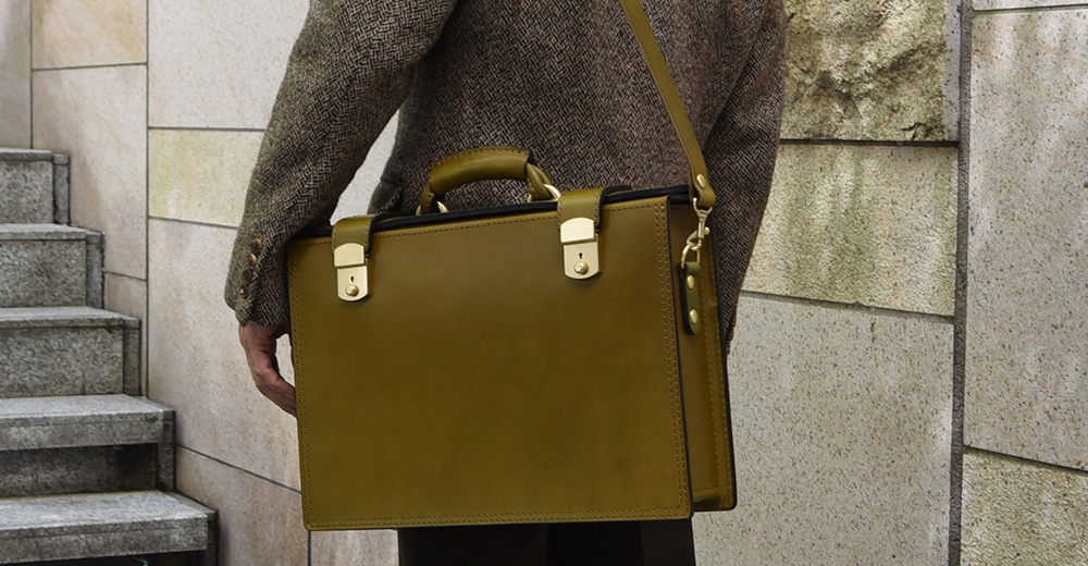 橄欖綠皮革製成的職人手作兩用機師箱，配有金屬鎖扣與提把，可肩背或手提。