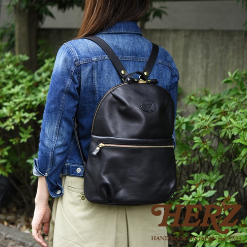 黑色皮革大開口圓角後背包，配有金色拉鍊和品牌標誌，由一位身穿牛仔夾克和卡其色長褲的女士背著。