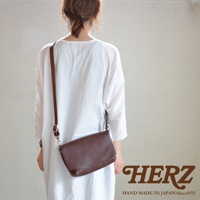 一名女子背對鏡頭，身穿白色寬鬆上衣，斜挎一個棕色皮革翻蓋斜背包，包包上有金屬扣環，右下角有HERZ品牌標誌和Hand Made in Japan Since 1973字樣。