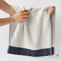 一條淺灰色、帶有細緻織紋的長纖有機棉斜紋浴巾，底部滾邊為深藍色斜紋布料，邊緣有細條紋裝飾。