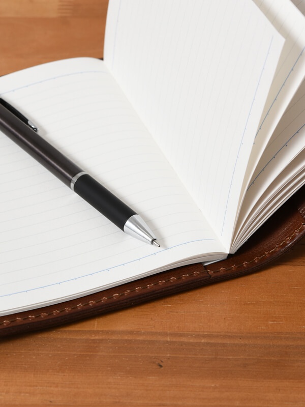 職人手作皮革A6記事本書套，深棕色皮革外殼，內含一本橫線筆記本，筆記本左頁放置一支黑色金屬筆。