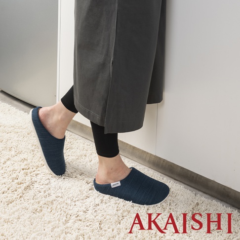 一雙深藍色、織紋布料的減壓包覆室內拖鞋，鞋側有白色小標籤，搭配黑色內搭褲和灰色長裙，站在白色絨毛地毯上。