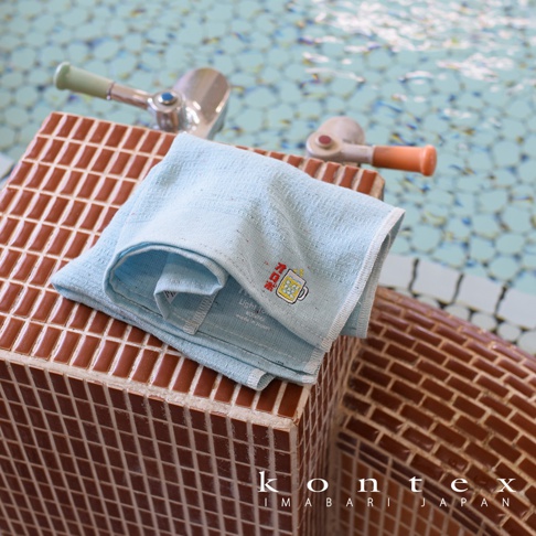 淺藍色薄輕長精繡桑拿毛巾，質地輕柔，帶有啤酒杯圖案繡花，放置於褐色馬賽克磚塊上，背景為水池。