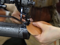 職人正在使用工業縫紉機，將一片淺棕色光滑皮革縫製成零錢包，皮革邊緣有細緻的縫線。