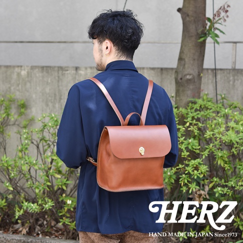 男士身穿深藍色上衣，背著一個棕色皮革翻蓋迷你後背包，背包採用簡約設計，配有金色鎖扣。