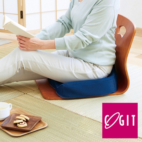 深藍色網狀材質的W型機能紓壓美尻坐墊，置於淺棕色木製椅凳上，一名女性穿著淺藍色上衣和白色長褲，正坐在坐墊上閱讀。