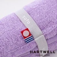 一條捲起來的淺紫色毛巾，上面繫著一條銀灰色織帶，織帶上印有 soto 字樣，織帶旁有一個紅底白圓點搭配藍白條紋的小標籤，毛巾的材質為柔軟的毛圈。