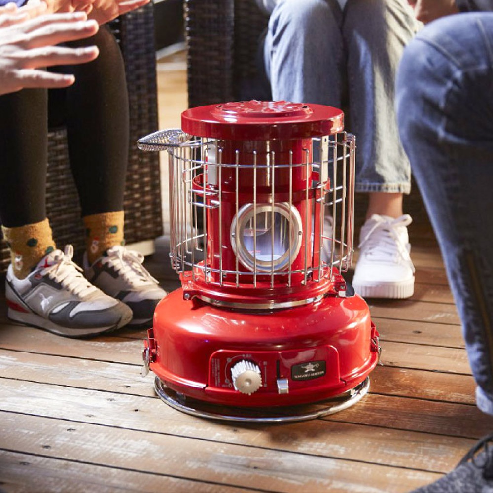 紅色的卡式瓦斯暖爐，設有金屬網罩和圓形觀察窗，底部有白色旋鈕，置於木質地板上，周圍有幾個人。