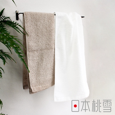 兩條Furry雙股紗絨毛浴巾，一條為淺褐色，另一條為白色，皆懸掛於黑色毛巾架上，背景為白色牆面，左側有綠色植物葉片。