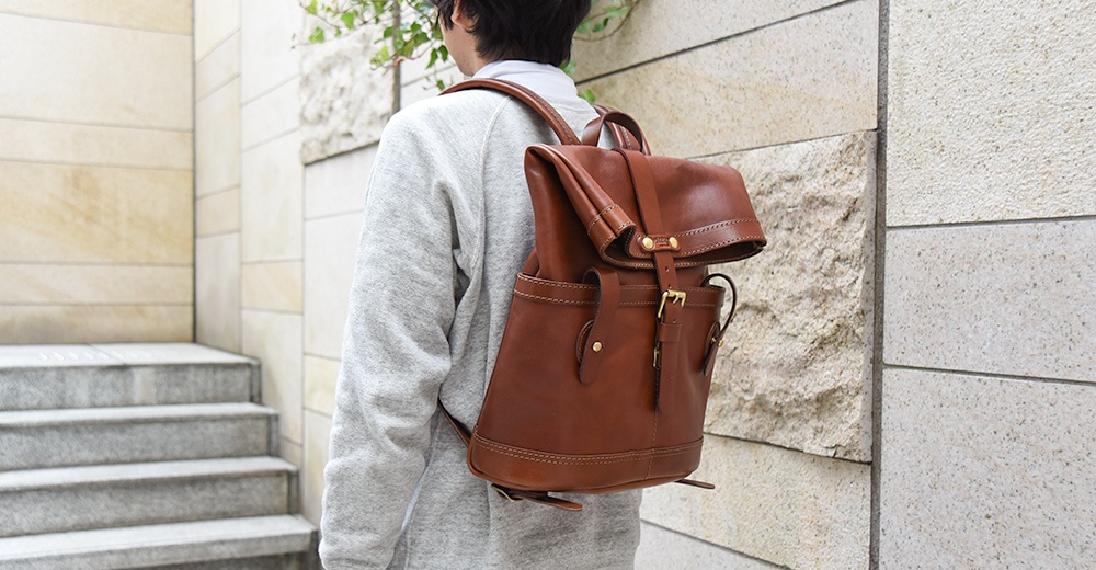 職人手作皮革雙口袋折蓋後背包，呈現溫潤的咖啡色調，質感皮革搭配金屬釦件，設計簡約實用。