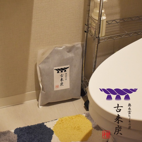 灰色包裝袋裝有「古來炭」字樣和繩結圖案，旁邊為白色馬桶，上方置物架上有白色瓶裝物。