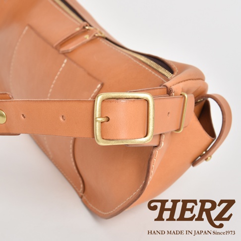 HERZ 職人手作皮革大容量斜背包，特寫金屬釦環與皮革揹帶的細節，下方標示 HERZ HAND MADE IN JAPAN Since1973