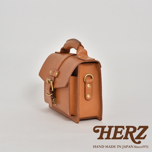 職人手作的經典混合風迷你單肩包，採用天然皮革材質，呈現溫潤的棕褐色。包款設計簡潔，帶有頂部提把與可調節的側背帶，並配有復古風格的銅色金屬扣環與铆釘，展現精緻工藝。