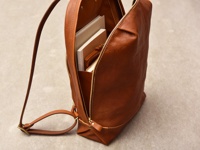 一個簡約褶皺風的棕色皮革後背包，部分拉鍊敞開，露出內部裝有幾本書籍。背包有一條皮革肩帶。