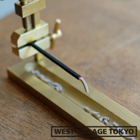 黃銅夾式水平線香架，夾持一根燃燒中的黑色線香，線香末端呈現紅熱狀態，並有細微的煙霧飄出。線香架為金屬材質，表面光滑，呈現黃銅色澤，底部有收集香灰的槽狀設計。