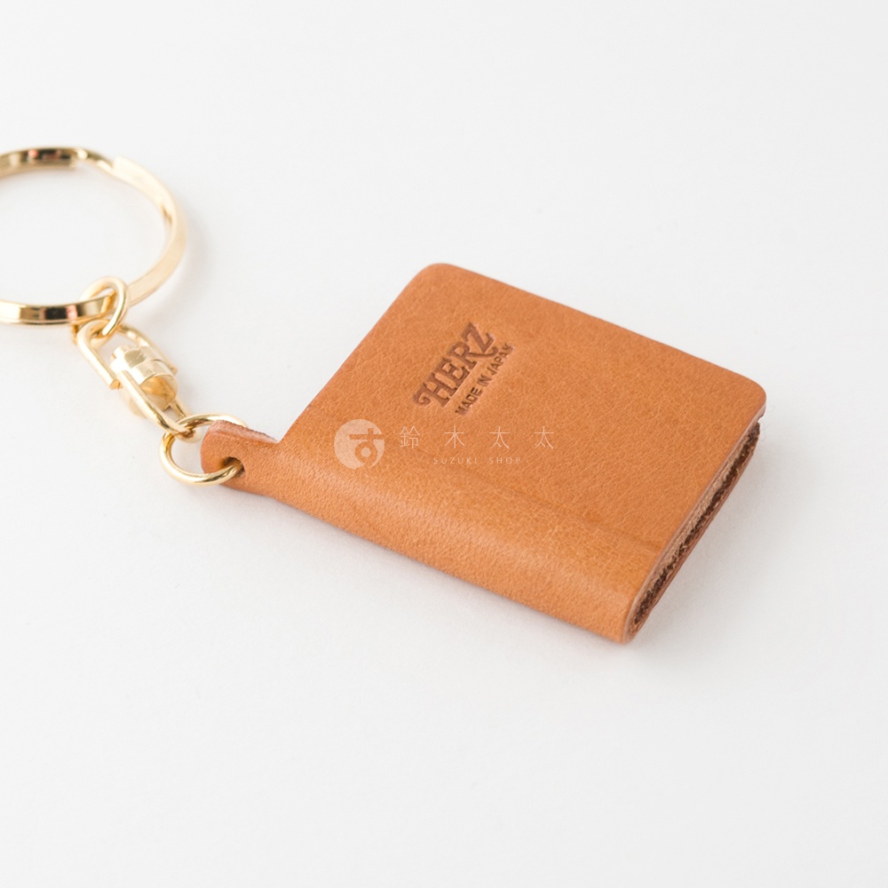 職人手作皮革書本造型鑰匙圈，採用棕褐色皮革，搭配金色金屬扣環與掛鍊，皮革表面壓印有「HERZ」及「MADE IN JAPAN」字樣。