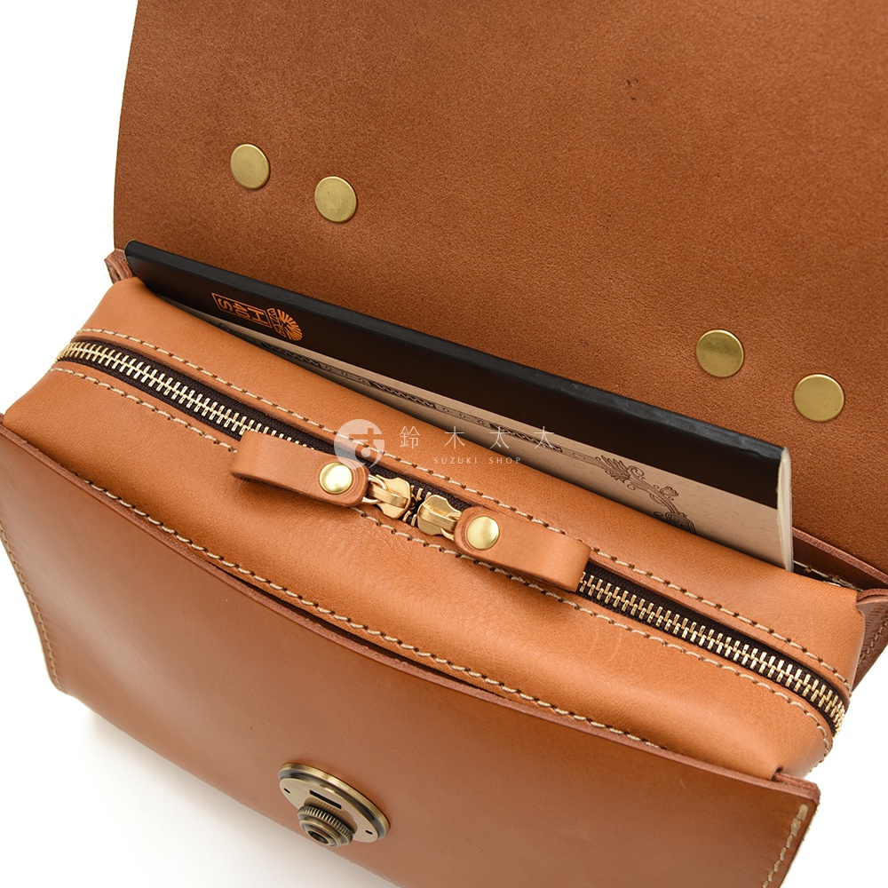 駝色皮革多功能收納單肩包，內部拉鍊夾層內放著一本日文書，包包上方有三個金屬卯扣。
