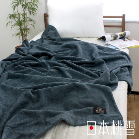 深藍色今治飯店毛巾被，材質柔軟，覆蓋在白色床單上，一旁有雜誌和綠植，角落有品牌標籤。