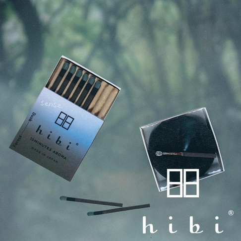 這張照片呈現了 hibi 香氛火柴禮盒，左側為一盒打開的香氛火柴，盒身印有 hibi 品牌標誌與 Dusk Forest 字樣；右側是一個圓形底座，上面放置著一根正在燃燒發出煙霧的火柴，下方還有兩根散落的火柴，背景呈現柔焦的森林意境。