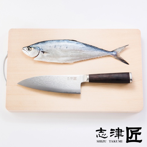木質砧板上擺放著一條銀灰色魚和一把 Santoku 萬用刀。刀刃採用大馬士革鋼材質，呈現獨特紋路，手柄為深棕色木質。