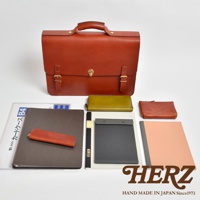 棕色皮革公事包，配有金色鎖扣和搭扣。包旁擺放著綠色長夾、棕色零錢包、灰色文件夾、筆和粉色筆記本。